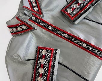 May include: A silver-gray garment with decorative embellishments. The garment features red, black, and silver geometric patterns and sparkling accents. The edges are trimmed with black beading. The fabric has a subtle sheen.