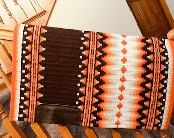 May include: A rectangular horse saddle pad featuring a thick, dark brown center panel flanked by vertical bands of orange, white, and black geometric patterns. The pad includes a dark brown leather wear patch at the bottom corner.