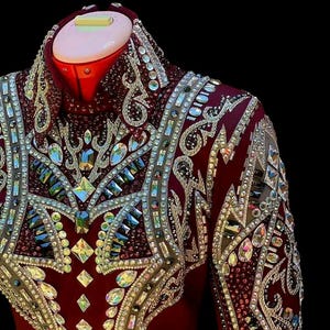 May include: A burgundy equestrian show jacket with intricate silver and crystal embellishments. The jacket features a high collar and is adorned with a dazzling array of rhinestones and gemstones, creating a luxurious and eye-catching design. The jacket is displayed on a mannequin.