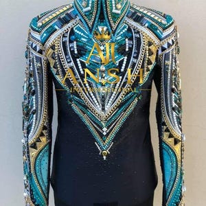 May include: A black long-sleeve shirt featuring a high collar and intricate embellishments in teal, gold, and white. The front is decorated with crystals, metallic chains, and geometric patterns. The text AJI INTERNATIONAL is printed in gold.