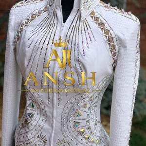 May include: A white long-sleeved show shirt featuring a high collar, a front zipper, and intricate crystal embellishments in geometric and linear patterns. The shoulders and upper sleeves include lace-up detailing with silver accents and rhinestones.