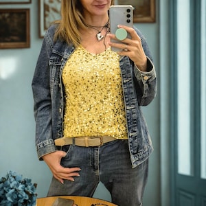 Op de afbeelding: Outfit met een spijkerjasje en jeans. Het jasje heeft een medium wassing en de jeans een donkerdere wassing. Een gele top met pailletten wordt onder het jasje gedragen. Een beige riem wordt bij de jeans gedragen. Een zilveren hartketting is ook zichtbaar.