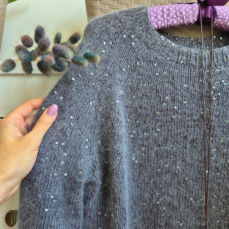 May include: A gray knit sweater with a crew neck and scattered sequins. The sweater has a soft, textured appearance. The image shows a close-up of the shoulder and sleeve of the garment.