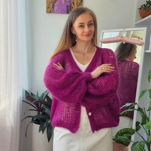 Può includere: Un cardigan color magenta vivace con scollo a V, caratterizzato da una trama a maglia larga, maniche lunghe voluminose e chiusura frontale con bottoni. Il capo è indossato sopra un top bianco semplice, con una vestibilità ampia e soffice.