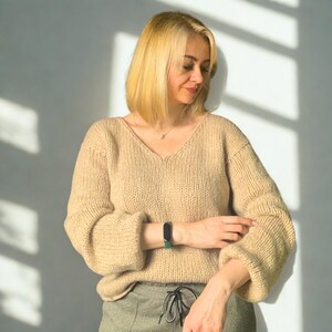Puede incluir: Un su&eacute;ter de punto beige de manga larga con cuello en V y mangas holgadas. El su&eacute;ter presenta un patr&oacute;n de punto grueso y texturizado, combinado con pantalones verde oliva con cintura de cord&oacute;n y un rastreador de actividad f&iacute;sico negro en la mu&ntilde;eca.