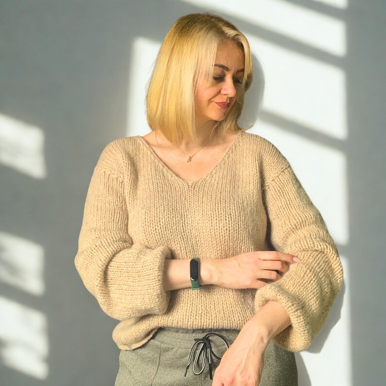 Puede incluir: Un su&eacute;ter de punto beige de manga larga con cuello en V y mangas holgadas. El su&eacute;ter presenta un patr&oacute;n de punto grueso y texturizado, combinado con pantalones verde oliva con cintura de cord&oacute;n y un rastreador de actividad f&iacute;sico negro en la mu&ntilde;eca.