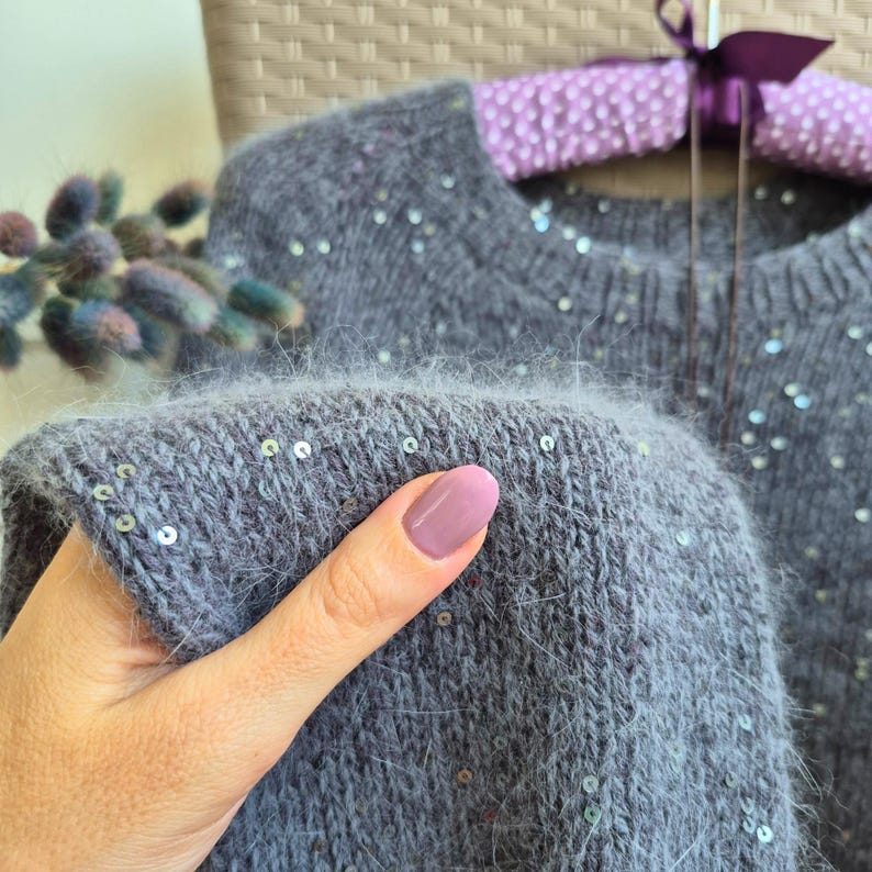May include: A gray sweater with scattered sequins. The soft knitwear is held up by a hand with a pink nail polish. The sweater is on a purple hanger with a ribbon.