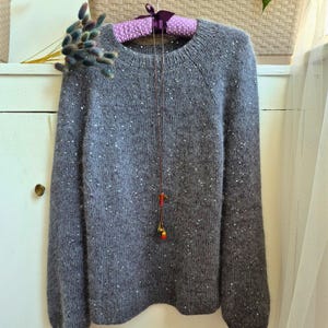 May include: A gray, long-sleeved sweater with a crew neck and scattered white sequins. The sweater is hanging on a polka-dot padded hanger. A delicate necklace with red and gold beads hangs in front of the sweater.