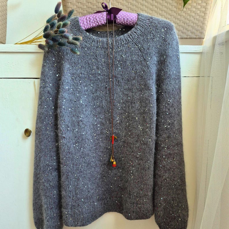 May include: A gray, long-sleeved sweater with a crew neck and scattered white sequins. The sweater is hanging on a polka-dot padded hanger. A delicate necklace with red and gold beads hangs in front of the sweater.