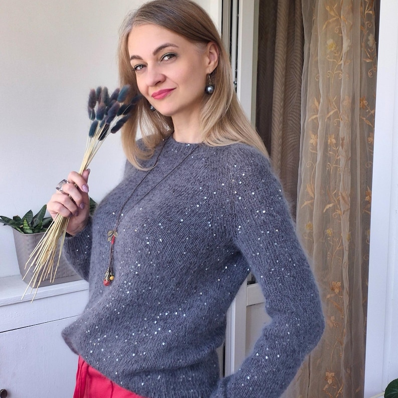 May include: A gray sweater with scattered white sequins. The long-sleeved knitwear has a crew neck. The person is holding a bouquet of dried flowers. The person is wearing a long necklace and red pants.