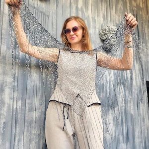May include: A woman wearing a sleeveless top and pants. She is holding up a sheer, black, beaded shawl. The top is white with a black pattern and trim. The pants are a light beige color. The background is a gray wooden wall.