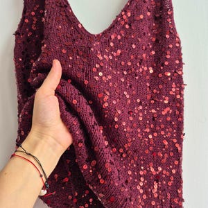 May include: A burgundy sequined vest top with a V-neck. The top is made of a knitted fabric and is covered in small, round, red sequins. The straps are thin, and the overall style is casual.