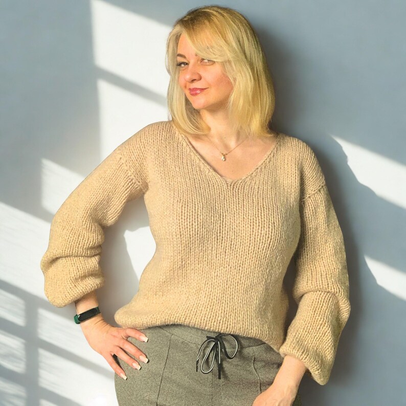 Puede incluir: Un su&eacute;ter beige de manga larga con cuello en V y textura de punto grueso, combinado con pantalones grises con cintura de cord&oacute;n. El su&eacute;ter tiene mangas ligeramente abullonadas y un ajuste relajado, mientras que los pantalones tienen un patr&oacute;n sutil.