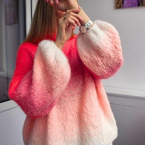 Puede incluir: Un suéter vibrante y de gran tamaño en un degradado de rosa y blanco. El suéter presenta mangas abullonadas y un cuello redondo. La parte superior es de un rosa coral brillante, que transiciona a un rosa más claro y blanco en las mangas y la parte inferior.
