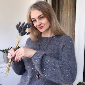 May include: A gray sweater with sequin embellishments. The sweater has long sleeves and a round neckline. The person in the image is holding a bouquet of dried flowers. A long necklace with colorful beads is also visible.