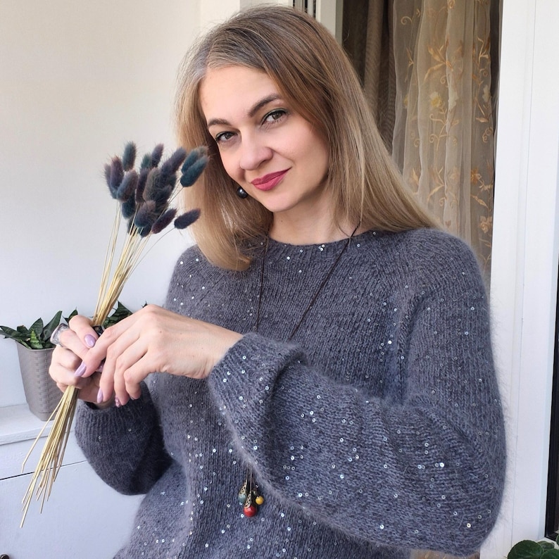 May include: A gray sweater with sequin embellishments. The sweater has long sleeves and a round neckline. The person in the image is holding a bouquet of dried flowers. A long necklace with colorful beads is also visible.