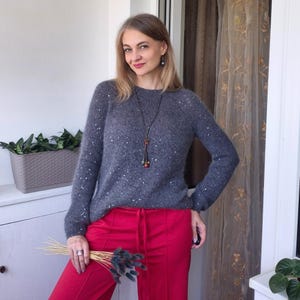 May include: A woman wearing a gray sweater with sparkling embellishments and red pants. The sweater has long sleeves and a round neckline. She is also wearing a long necklace with decorative beads. The pants have a drawstring waist.