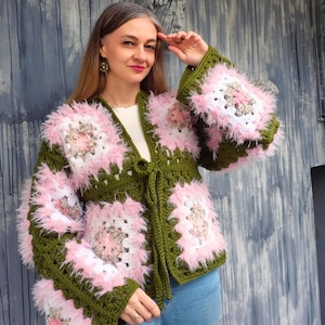 May include: A long-sleeved crochet cardigan featuring a pattern of olive green squares interspersed with fuzzy pink and white square motifs. The garment has a tie-front closure, a V-neckline, and a textured, shaggy appearance created by the yarn.