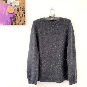 May include: A gray speckled sweater with a crew neck, hanging on a polka dot hanger. The sweater has long sleeves and a ribbed hem and cuffs. The background includes a painting with purple and gold tones.