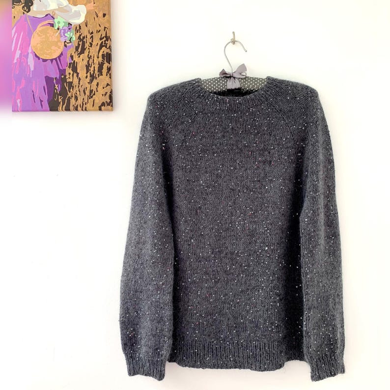 May include: A gray speckled sweater with a crew neck, hanging on a polka dot hanger. The sweater has long sleeves and a ribbed hem and cuffs. The background includes a painting with purple and gold tones.