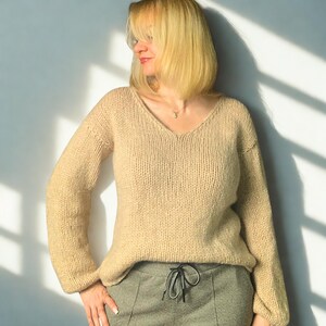 Puede incluir: Un su&eacute;ter beige de manga larga con cuello en V, confeccionado en tejido de punto grueso. El su&eacute;ter presenta un corte relajado con hombros ligeramente ca&iacute;dos y pu&ntilde;os acanalados. Se combina con pantalones grises con cierre de cord&oacute;n ajustable.