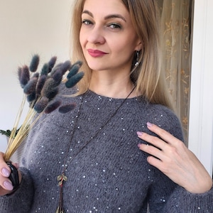 May include: A gray sweater with sequin embellishments. The sweater is paired with a long necklace with leaf pendants. The person is holding a bouquet of dried flowers with blue and green accents.