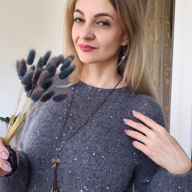 May include: A gray sweater with sequin embellishments. The sweater is paired with a long necklace with leaf pendants. The person is holding a bouquet of dried flowers with blue and green accents.