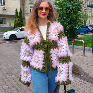Op de afbeelding: Handgemaakte gehaakte jas met een granny square ontwerp. De jas combineert olijfgroen, wit en roze garen, met pluizige roze accenten. Het heeft een striksluiting in de taille en wordt gedragen met een blauwe jeans.