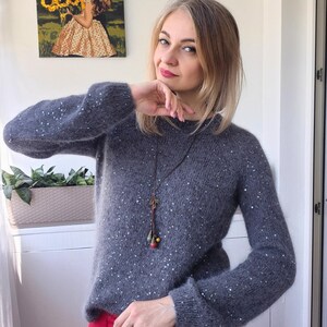 May include: A gray sweater with white sequins is shown. The sweater has long sleeves and a round neckline. A long necklace with colorful beads hangs down the front. The person is wearing red pants.