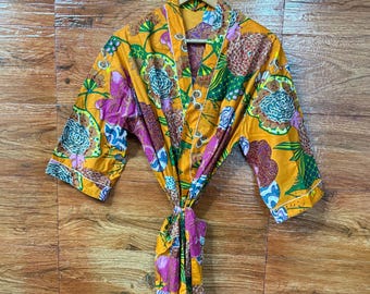Puede incluir: Una bata vibrante con estampado floral en tonos amarillos, rosas, verdes y marrones, colgada de una percha de madera. La bata presenta un cierre de lazo y un diseño botánico colorido, perfecta para descansar o como una elegante prenda.