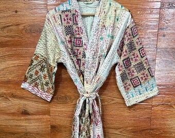 Puede incluir: Bata color crema con un estampado geométrico en tonos marrón, rosa y azul. La bata tiene un cierre de lazo y está colgada de una percha de madera. La tela parece acolchada o patchwork.