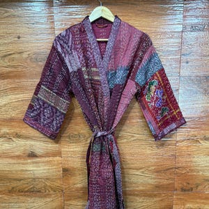 Peut inclure: Une robe patchwork dans les tons bordeaux, violet et gris, avec une ceinture assortie. La robe présente un col en V et des manches trois-quarts. Le tissu a une texture matelassée avec des coutures visibles, suspendue à un cintre en bois.