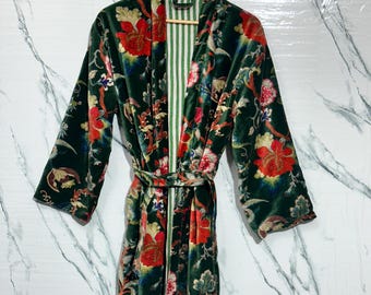 May include: A long, dark green velvet robe with a floral pattern in red, yellow, and blue. The robe has long sleeves and a matching belt. The interior lining is striped with green and white. The robe is hanging on a wooden hanger.