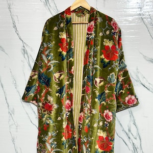 Könnte beinhalten: Olivgrüner Samt-Kimono mit floralem Muster aus roten und rosa Blüten, blauen Blättern und goldenen Akzenten. Das offene Kleidungsstück hängt an einem Holzbügel und zeigt ein gestreiftes Innenfutter.