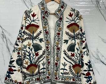 May include: Off-white jacket with intricate floral embroidery in shades of red, green, blue, and gold. The jacket features a mandarin collar and is displayed on a wooden hanger against a marble-patterned background. The embroidery details are visible on the front, sleeves, and collar.