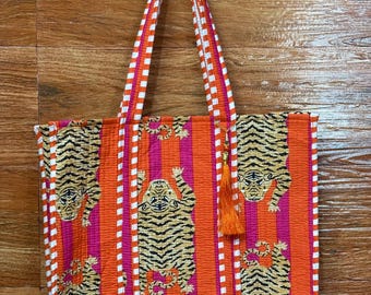 Könnte beinhalten: Eine lebendige Tragetasche mit Tigerdruck. Die Tasche hat orange, rosa und weiße vertikale Streifen mit Tigerillustrationen. Die Griffe sind orange und weiß gestreift. Eine orangefarbene Quaste hängt an der Seite.