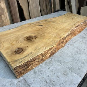 May include: A rectangular, unfinished wooden slab with a natural edge. The light-colored wood features visible grain patterns and two prominent knots. The slab is likely intended for use as a tabletop or similar woodworking project.