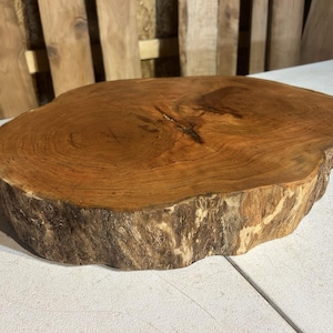 May include: A round, natural wood slab with a rich brown color and visible grain patterns. The edges of the slab have a rough, bark-like texture. This rustic piece could be used as a serving board or decorative accent.