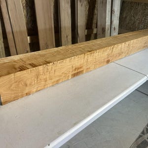 May include: A long, rectangular wooden beam with a smooth, polished finish. The wood features a light golden-brown color with visible natural grain patterns, knots, and subtle variations in texture throughout the length of the solid timber piece.