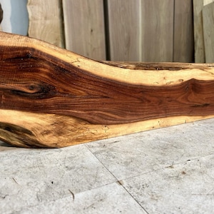 May include: A rectangular piece of natural wood with a live edge, featuring a dark brown heartwood center and lighter tan sapwood along the top and bottom edges. The wood grain is visible throughout, showcasing organic patterns and textures.