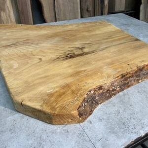 May include: A natural wood cutting board with a live edge. The board is rectangular with rounded corners and a light brown color. The live edge has a rough, textured appearance.