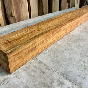May include: A long, rectangular wooden beam with a natural honey-brown finish. The beam displays a prominent wood grain pattern with visible knots and variations in color, featuring a smooth, planed surface and sharp, clean edges along its length.