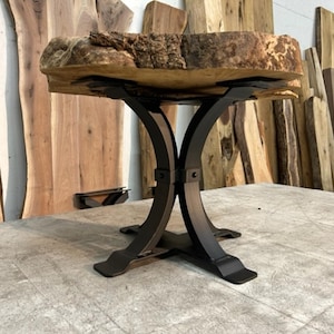 May include: A live edge wood table with a natural, rough-hewn top. The tabletop is supported by a black, curved metal base. The table is a unique piece of furniture, showcasing the natural beauty of the wood.