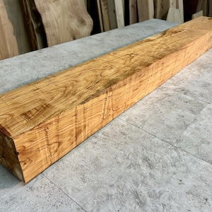 May include: A long, rectangular wooden beam featuring a natural light brown color with prominent, swirling dark grain patterns. The surface of the timber displays a smooth, polished finish that highlights the intricate, organic texture of the wood.