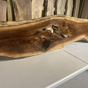 May include: A large, live edge wood slab with a rich, dark brown color and natural grain patterns. The slab has a unique shape with a large opening and is likely intended for use as a table or countertop. The wood appears to be walnut.