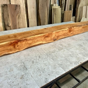May include: A long, rectangular, unfinished wooden beam with a natural edge. The wood has a warm, reddish-brown hue with lighter streaks and knots. It rests on a gray surface, with other wood pieces in the background. The beam is approximately 72 inches long.