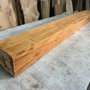 May include: A rectangular, solid wood beam with a natural, light brown finish. The beam has a rounded edge on one end and is displayed on a gray surface. The wood grain is visible, showcasing the natural texture and color variations.