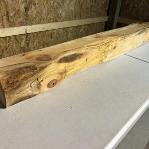 May include: A large, rectangular, unfinished wooden beam with a natural, light brown color and dark brown knots. The beam is lying horizontally on a white surface. The wood has a rough, textured appearance, suggesting it is for rustic decor.