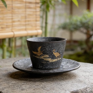 May include: A black ceramic teacup and saucer set with gold crane and water design. The teacup is cylindrical and sits on a matching saucer. The set is placed on a stone surface, with a bamboo blind and garden in the background.