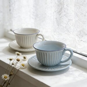 Puede incluir: Dos tazas de té y platillos de cerámica en crema y azul claro, con un diseño acanalado y un fino ribete azul. Las tazas están sobre un alféizar blanco con una cortina de encaje en el fondo. Unas pequeñas flores blancas en primer plano.